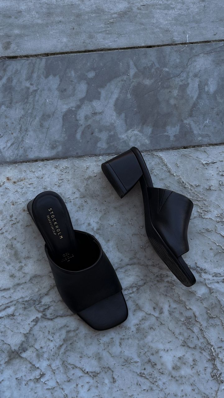 Svarta mules med blockklack och bred tårem i minimalistisk stil, placerade på ett marmorgolv.