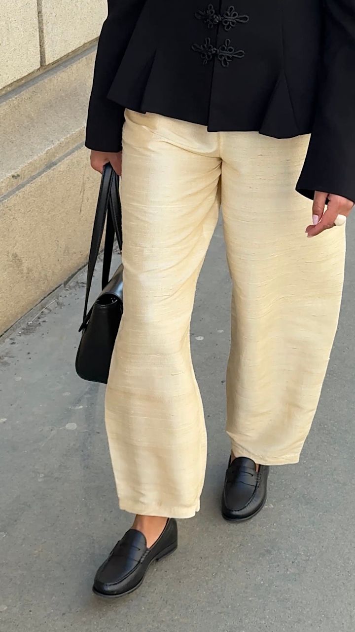 Person i svart jacka och beiga byxor bär klassiska svarta loafers i läder med en svart handväska.