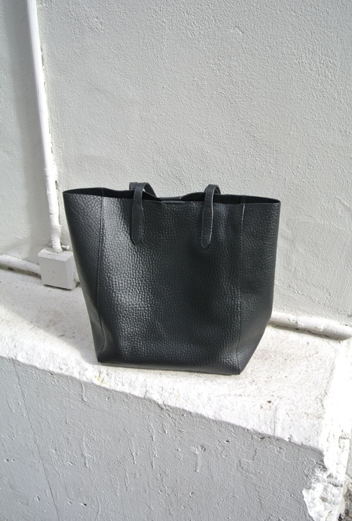 Svart skinnveske står på en vit murvägg med synliga rör och textur i bakgrunden.