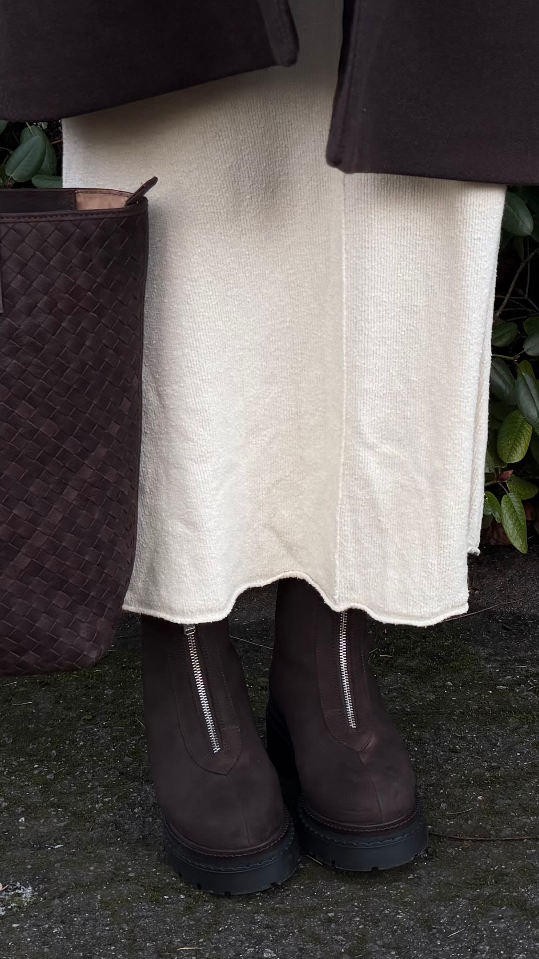 Bruna ankelkängor med dragkedja och kraftig sula under en krämvit kjol, bredvid en flätad brun väska på marken från Stockholm Design Group.