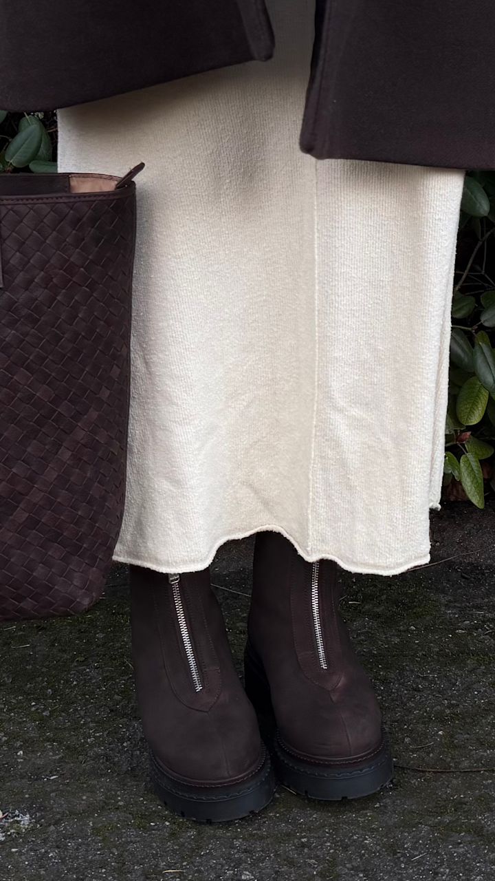 Bruna ankelkängor med dragkedja och kraftig sula under en krämvit kjol, bredvid en flätad brun väska på marken från Stockholm Design Group.