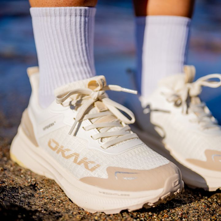 Hvite og beige løpesko fra Okaki med matchende hvite sokker, fotografert utendørs på steingrunn.