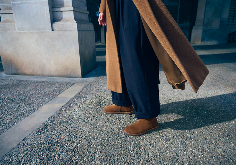 Person i mørkeblå bukser og kamelfarget frakk med brune semsket skinnsko i loafer-stil står på en steinbelagt overflate.