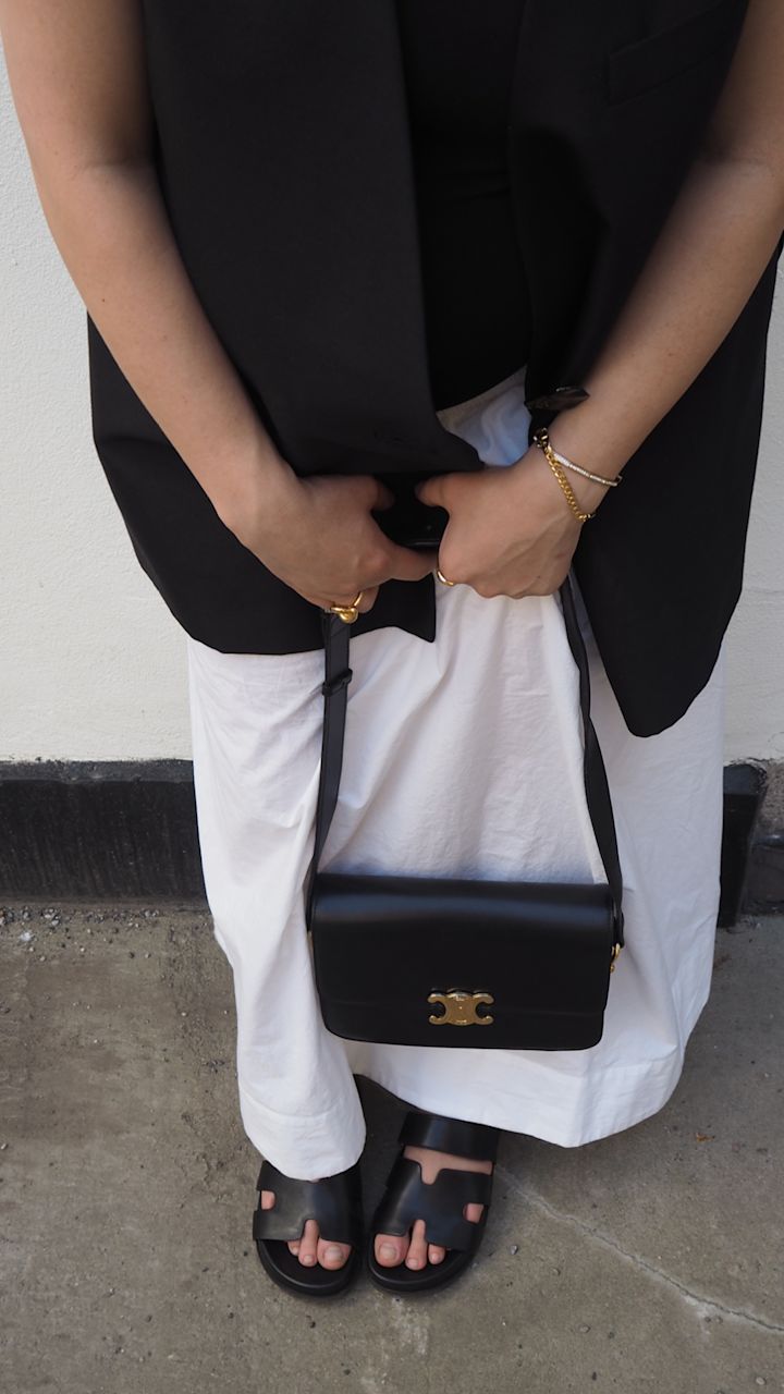 Personen håller en svart väska med guldfärgad spänne framtill, bär en vit lång kjol, svart väst och svarta sandaler, stående på betonggolv.