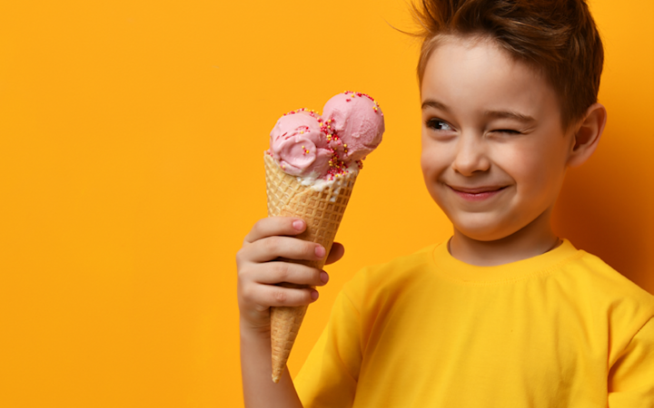 Pojke i gul t-shirt håller en glass med rosa kulor och strössel, och blinkar mot något utanför bilden mot en orange bakgrund.