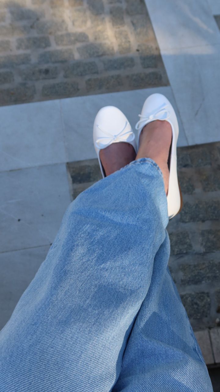 Hvite ballerinasko med små sløyfer fra Privé, brukt sammen med lyseblå jeans, sett ovenfra på steinbelagt underlag.