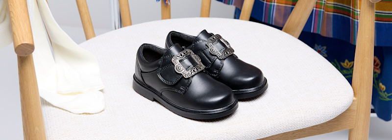 Svart lærsko med dekorativ metallspenne plassert på en hvit polstret stol med trearmstøtter.