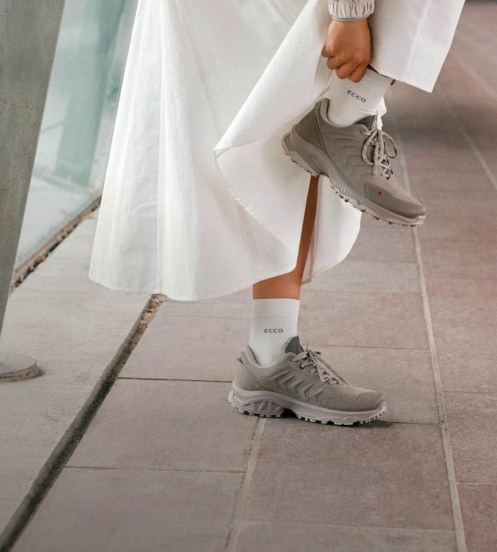 Kvinne i hvit kjole med grå ECCO-sneakers med robust såle og hvite ECCO-sokker, bøyer seg ned utendørs.