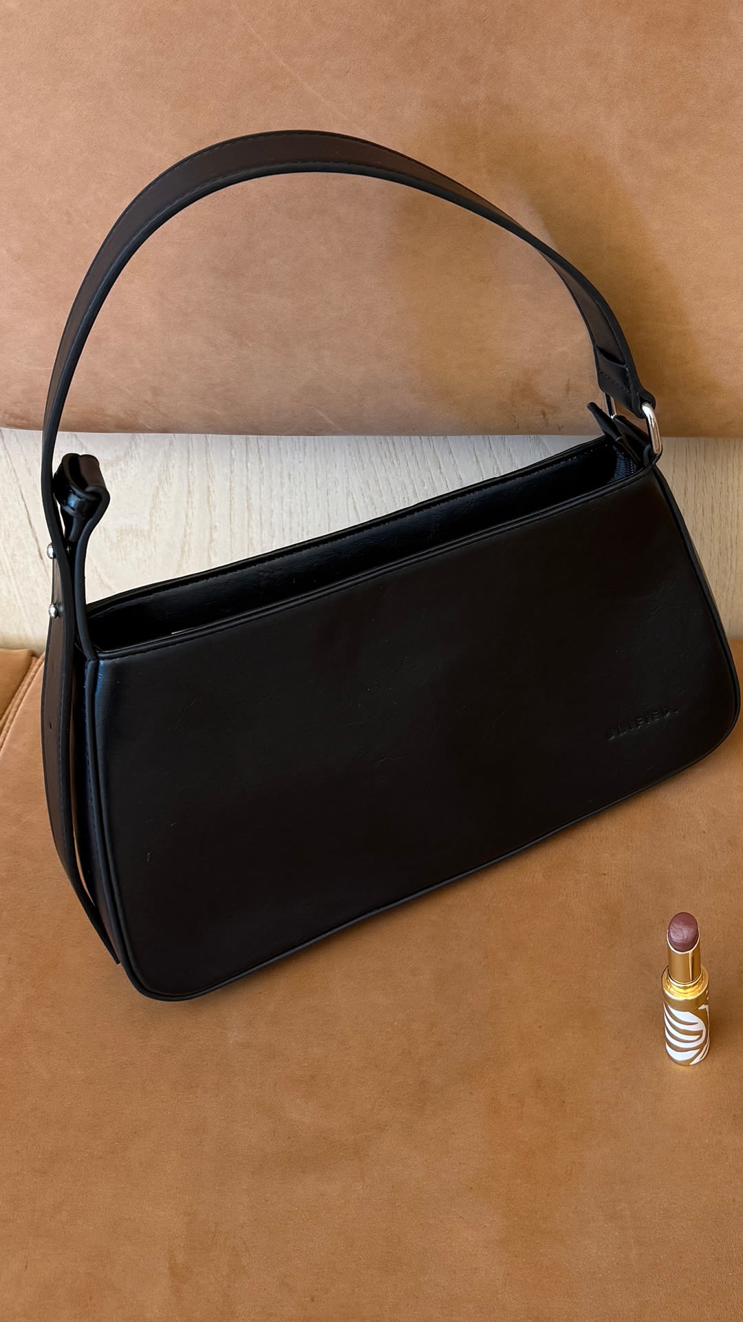Elegant svart handväska i klassisk design från Unified med handtag och spänne, placerad på brun bakgrund bredvid ett läppstift.