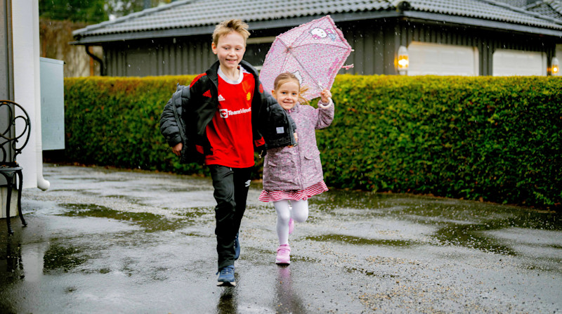To barn løper sammen på en våt asfaltvei i regnet, jenta holder et rosa paraply med Hello Kitty-motiv.