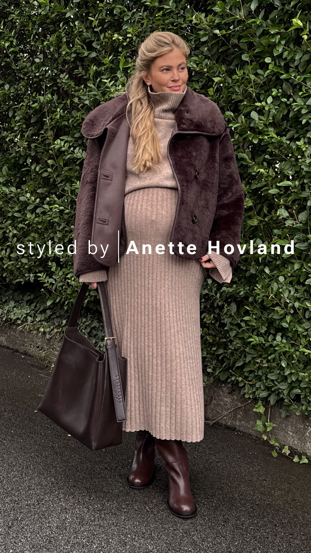 Influenser Anette Hovland i beige strikket antrekk med brun pelsjakke, matchende brun veske og elegante brune skinnstøvletter fra Sockholm Design Group foran grønn hekk.