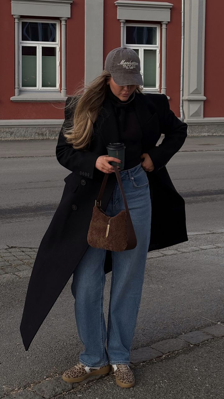 Kvinna bär en svart kappa, blå jeans, leopardmönstrade skor och en grå keps. Hon håller en kaffekopp och en brun pälsväska och står på trottoaren framför en byggnad med röda väggar och vita fönster.