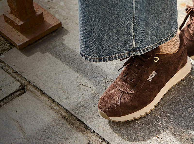 Brune semsket skinn sneakers med hvit såle og snøring, stylet med vide jeans på et steinunderlag.