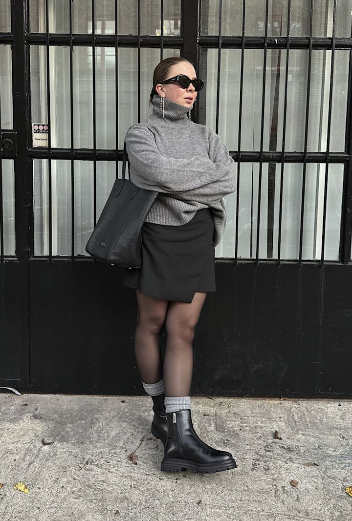 Kvinna bär en grå polotröja, svart kjol, svarta stövlar och solglasögon och står med armarna i kors framför en svart metallgrind.