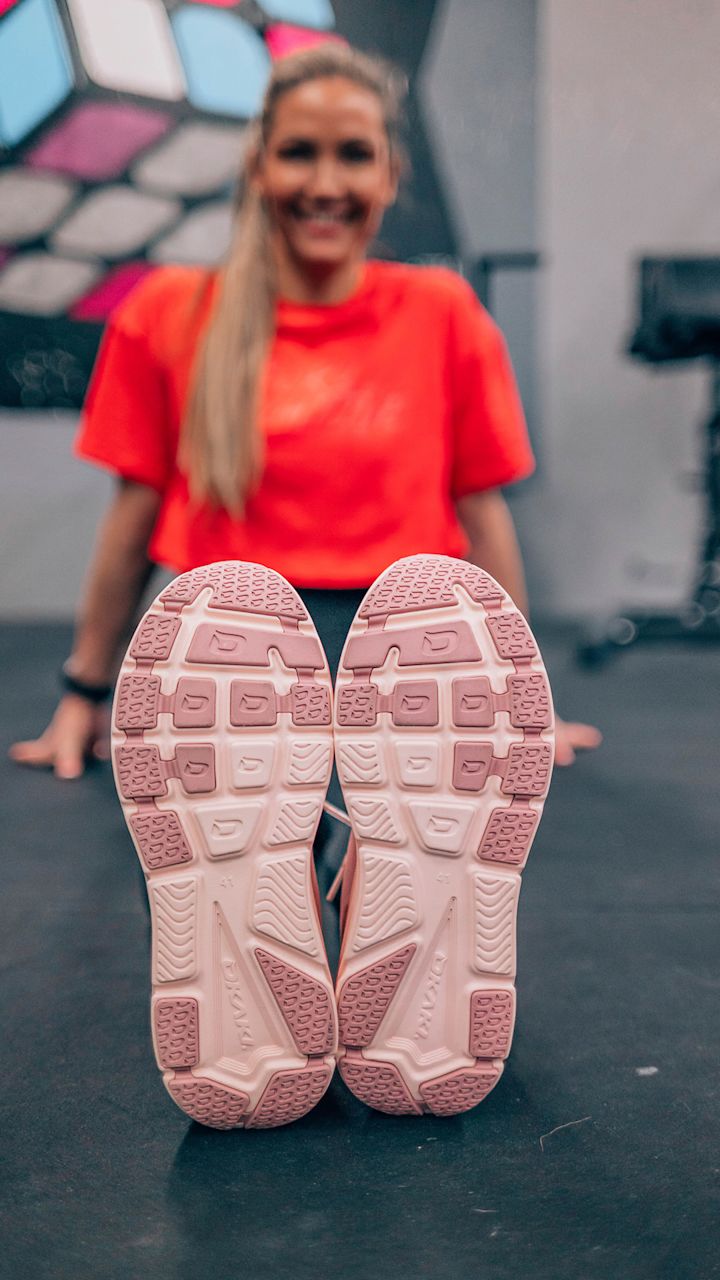 Kvinna i röd t-shirt sitter på golvet med fötterna utsträckta mot kameran, med fokus på undersidan av rosa joggingskor.