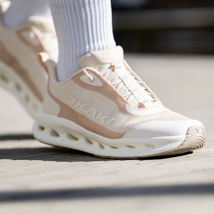 Beige og hvite sportssko med Okaki-logo, moderne design med perforert overflate og hvit såle, brukt med hvite sokker.