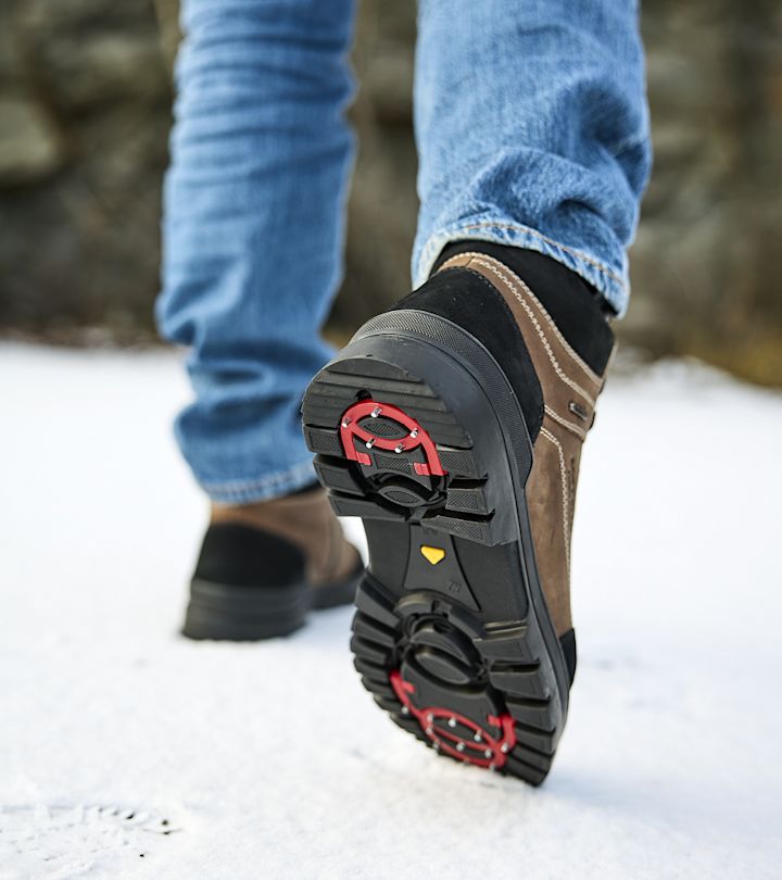 Skor med dubbar som går på snötäckt underlag, iförda blå jeans och bruna skor med svart sula.