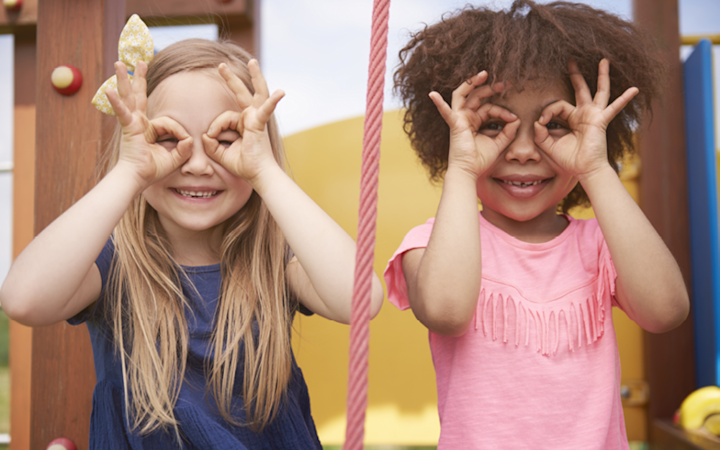 Två tjejer ler och formar glasögon med händerna framför ögonen på en lekplats.