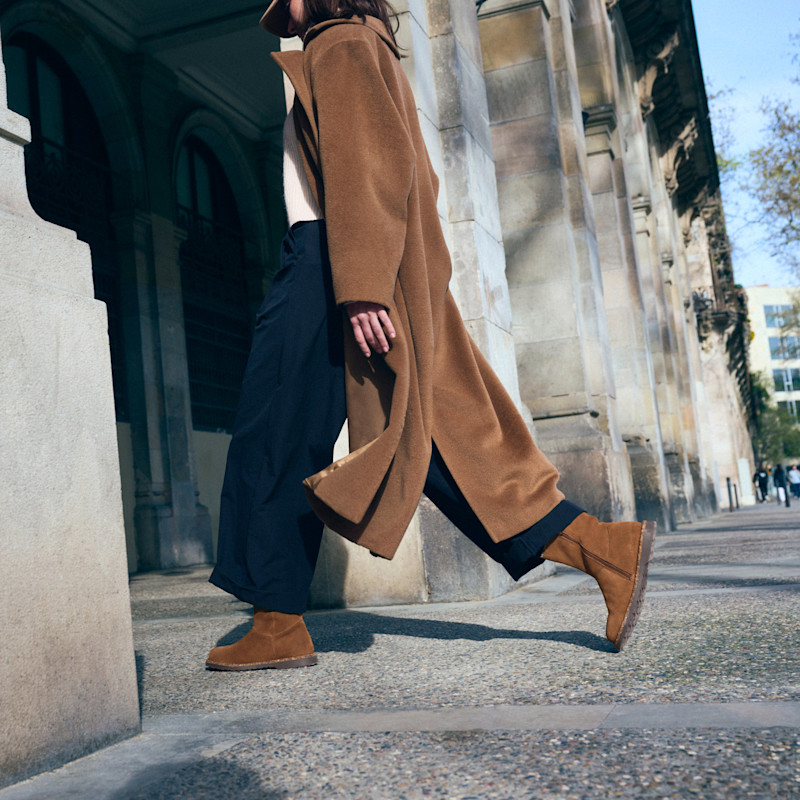 Person klädd i brun kappa och bruna stövlar som går på trottoaren vid en byggnad med stenfasad.