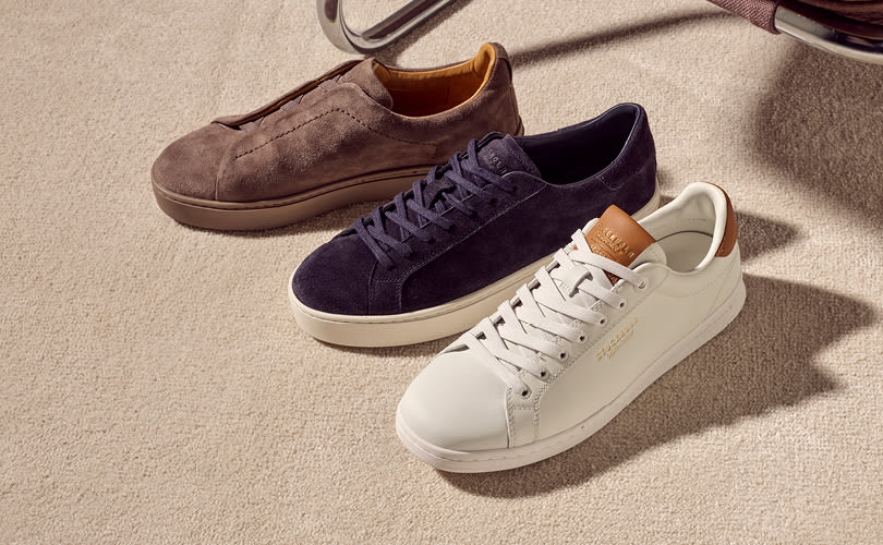 Tre elegante sneakers i ulike farger fra Stockholm Design Group: brun semsket, mørkeblå semsket og hvit med brune detaljer, plassert på beige underlag.