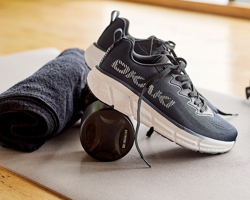 Svarte fritidssko fra Okaki med hvit såle ved siden av et håndkle, solbriller og en grønn treningsstrikk på en benk.