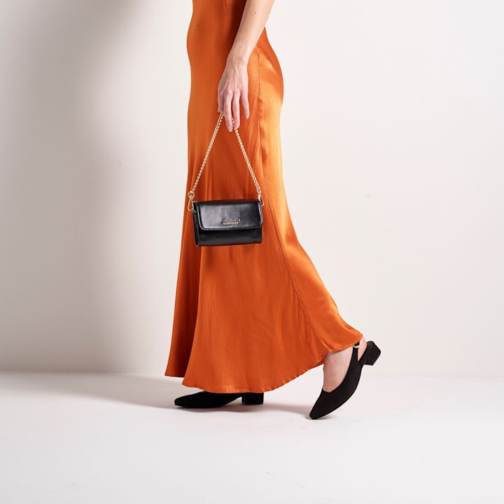 Person som går iført en lang, oransje kjole og svarte lave hæler, holder en liten svart veske med gullkjede i en hvit studio.