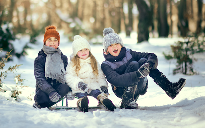 Tre barn klädda i vinterkläder sitter på en kälke i snön i en skog, med solljus som skiner genom träden.
