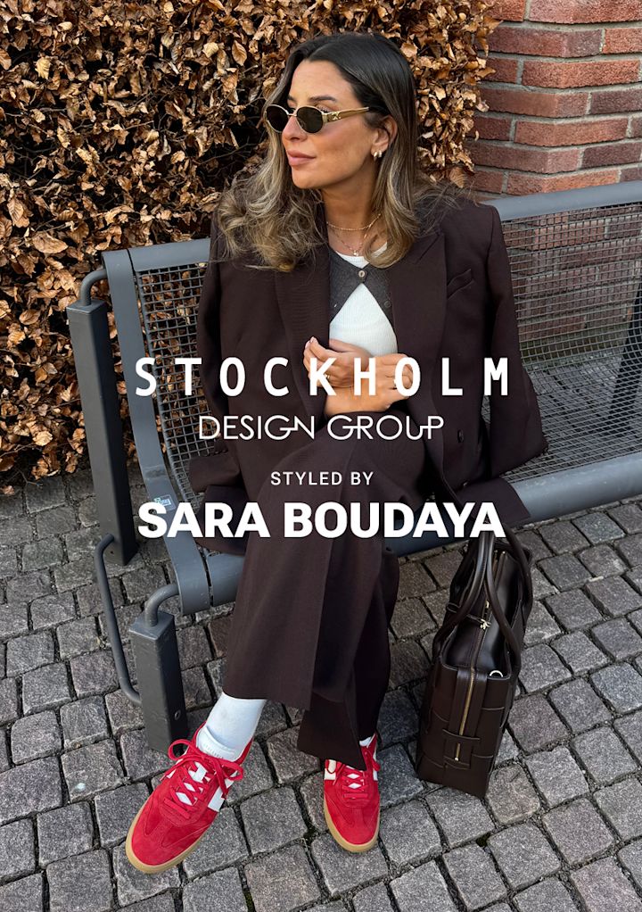 Kvinna med solglasögon och mörk kostym sitter på en bänk vid kullersten med en brun väska bredvid sig, framför torra löv och en tegelvägg. Text: "Stockholm Design Group styled by Sara Boudaya".