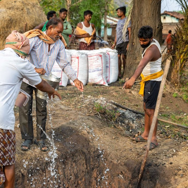 Män som häller vatten från hinkar ner i en utomhusdike, med flera män som står i bakgrunden nära stora säckar och träd.