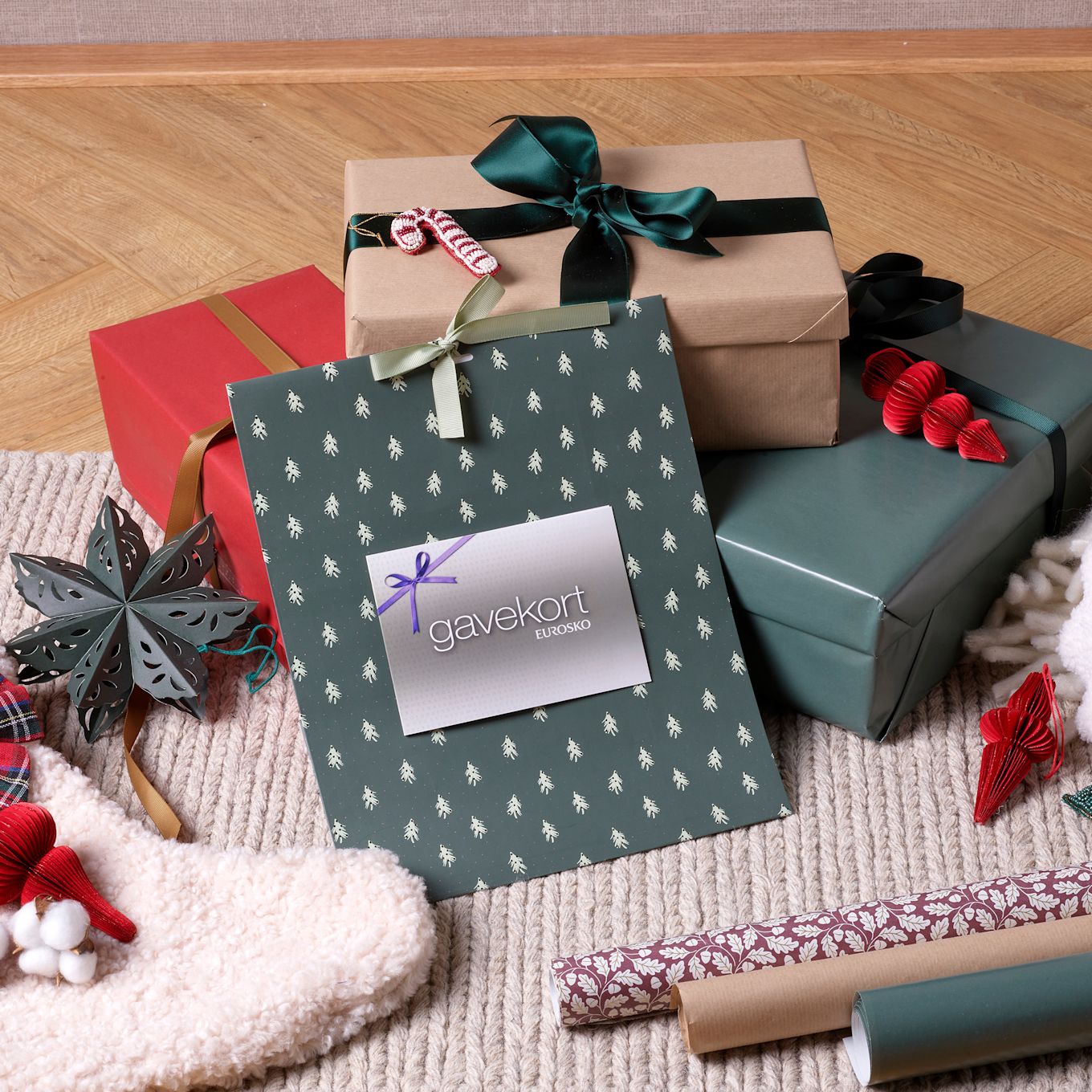 Julklappar i olika färger med ett grönt presentkort från Eurosko i centrum, omgivna av julpynt på en stickad matta.