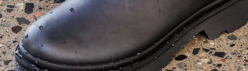 Vanntett svart støvel med vanndråper på overflaten, plassert på en steinlagt overflate.