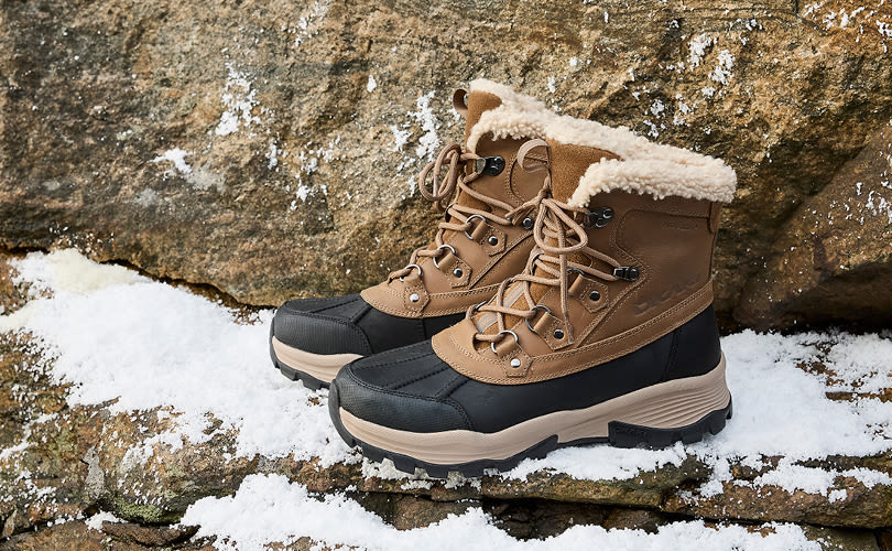 Ljusbruna och svarta vinterkängor med fluffig pälskant står på snötäckt klippa, robusta och stilfulla för vinterbruk.