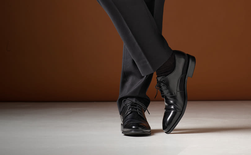 Elegante svarte skinnsko med snøring, brukt med mørk dress. Polert overflate og klassisk formell stil mot brun bakgrunn.