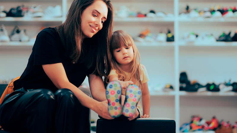 Kvinna hjälper ett litet barn att prova färgglada skor i en skobutik med hyllor fulla av skor i bakgrunden.