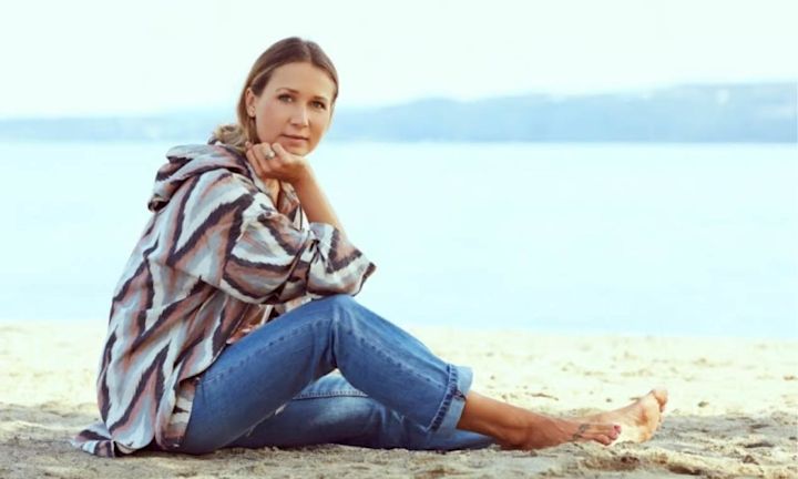 Kvinne sitter på sandstrand med sjø og fjell i bakgrunnen, iført mønstret genser og blå jeans med bare føtter.