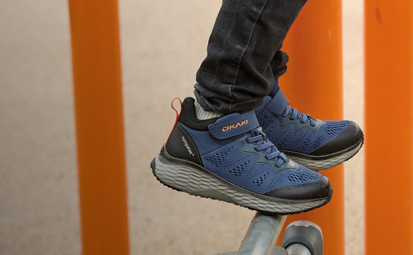 Blå och svarta OKAKI löparskor med ventilerad design och tjock grå sula, burna med mörka jeans mot orange bakgrund.