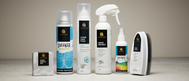 Flere Solitaire skopleieprodukter i ulike beholdere, inkludert Suede Nubuck Box, Sneaker Protector spray, Combi Care Foam, Water Protect spray, Sneaker Magic Cleaner og Shoe Shine børste, plassert på en grå overflate med nøytral bakgrunn.