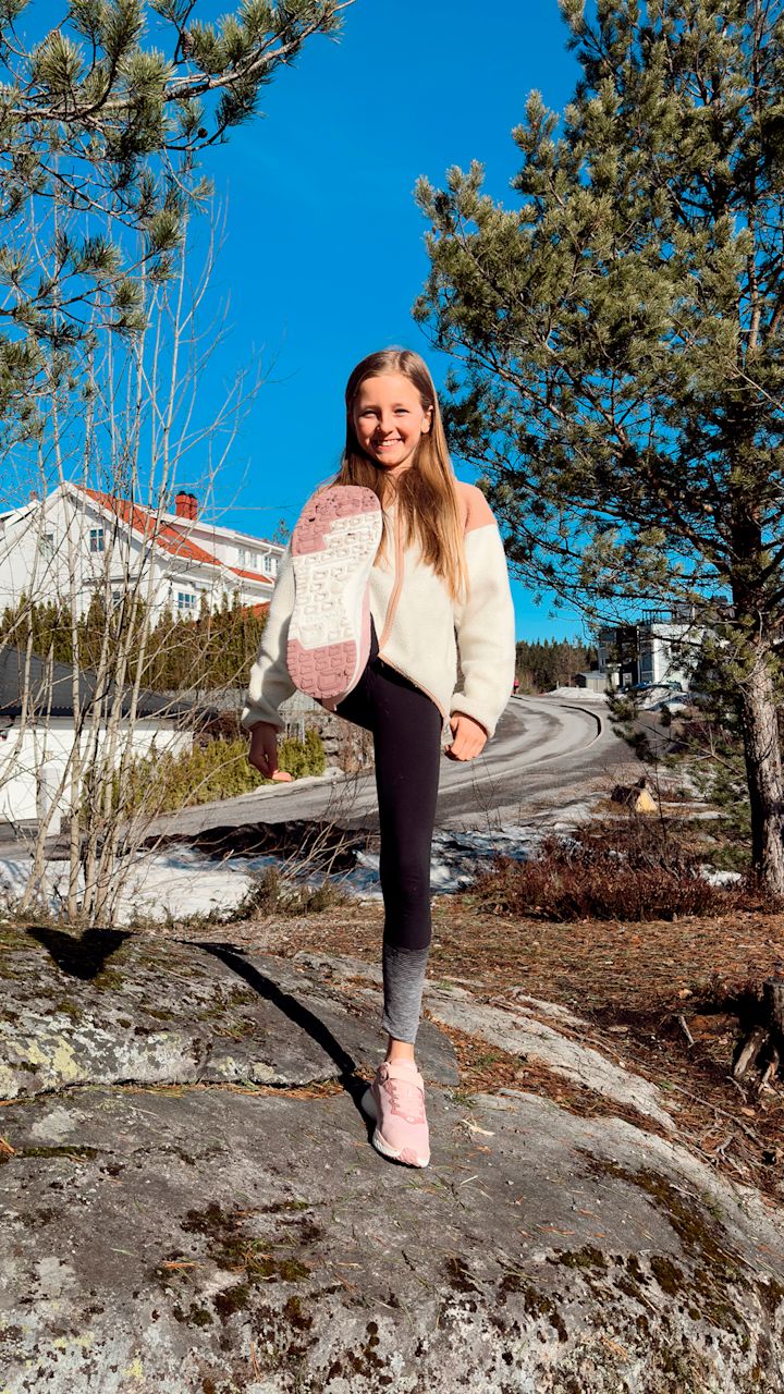 Jente i lys genser og svarte leggings løfter foten med rosa joggesko mot kameraet utendørs på en stein med trær og hus i bakgrunnen under blå himmel.