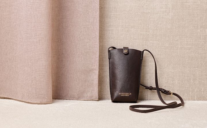 En liten brun läderväska med axelrem från Stockholm Design Group mot en beige bakgrund med textildraperier.