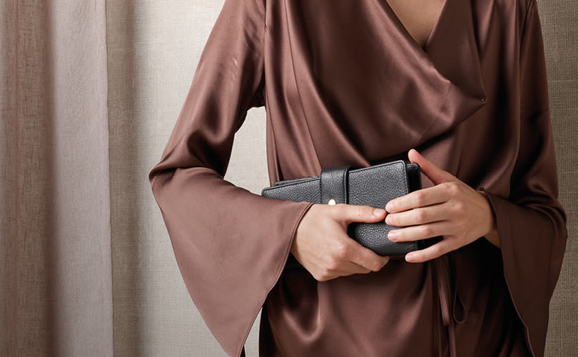 Person i brun sidenkimono håller en svart läderplånbok mot beige bakgrund.