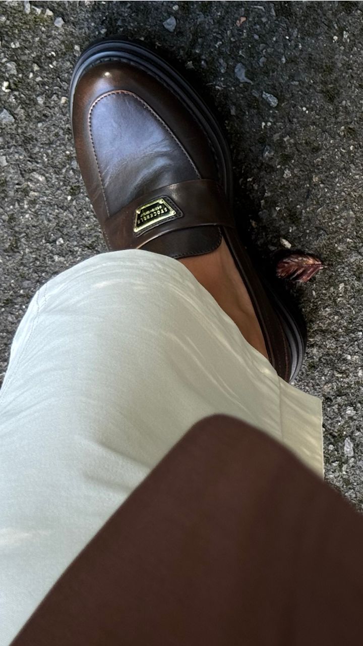 Elegant brun loafer från Stockholm Design Group med gulddetalj och svart sula, buren med vita byxor på asfalterad mark.