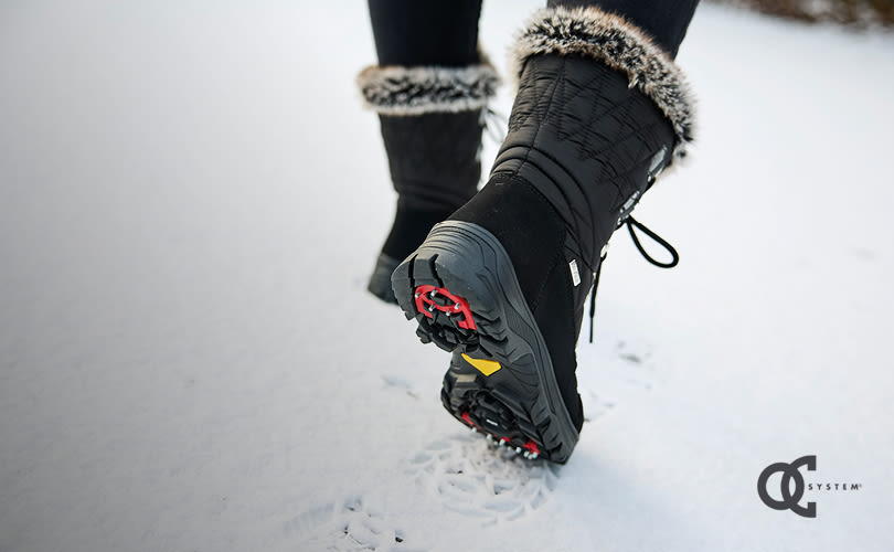 Svarta vinterkängor med pälskant och vändbara broddar som lämnar fotspår i snön.