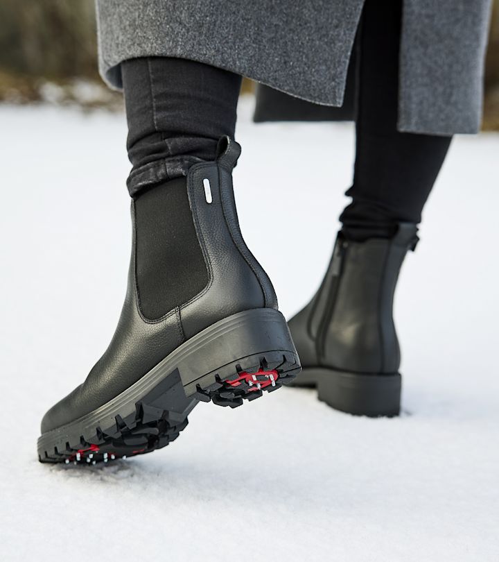 Svarta vinterstövlar med grovt mönster och röda dubbar på sulan, som går på snö under en grå kappa och svarta byxor.