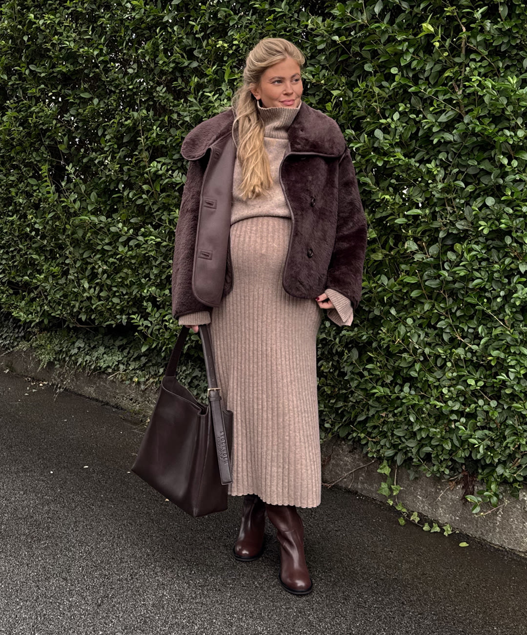 Person i beige strikket kjole og brun pelsjakke, med brune skinnstøvletter og matchende veske foran grønn hekk.