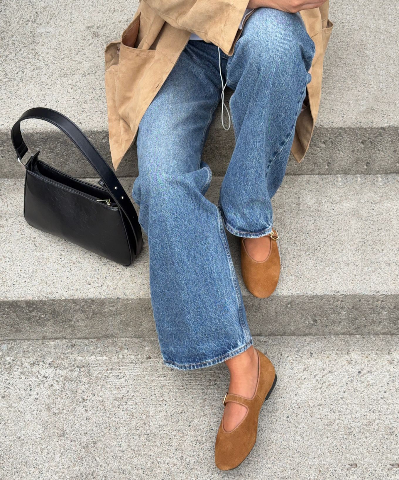 Person i beige frakk og jeans sitter på trapp med brune semsket Mary Jane-sko og svart skinnveske ved siden av.