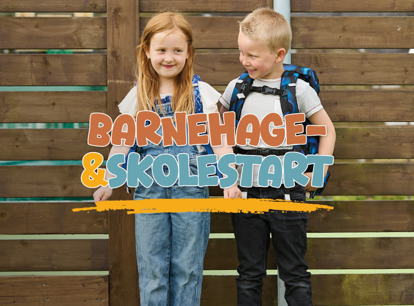 To barn smiler foran trevegg med teksten "BARNEHAGE- &SKOLESTART". Gutten har skolesekk og jenta har på seg jeans.
