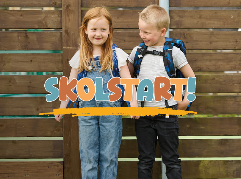Två barn framför trävägg med texten "SKOLSTART!". Flickan i jeansoverall och pojken med blå ryggsäck.
