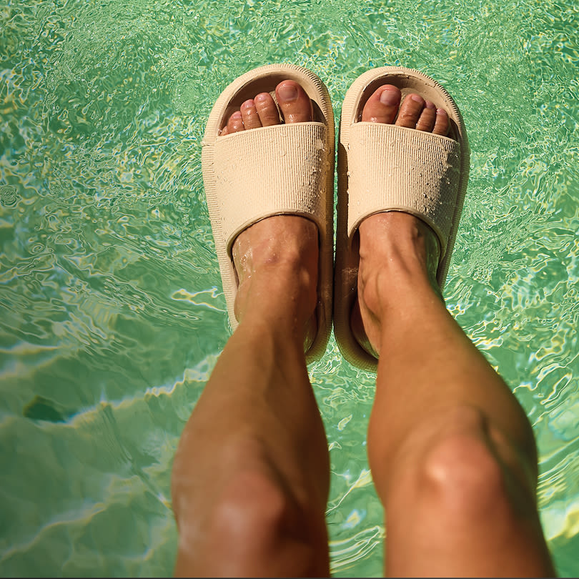 Beige slippers på føtter i klart, turkist bassengvann sett ovenfra.