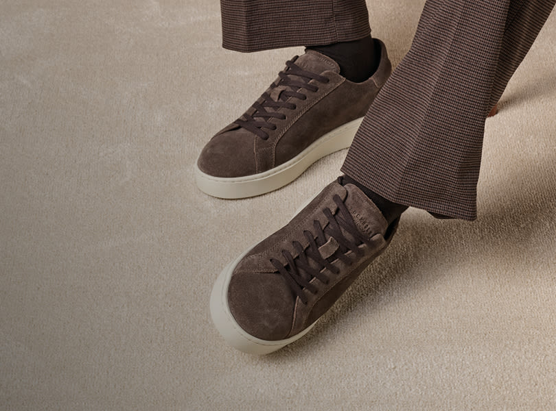 Bruna mocka sneakers från Stockholm Design Group med vita sulor, burna med rutiga byxor på en beige matta i elegant casual stil.
