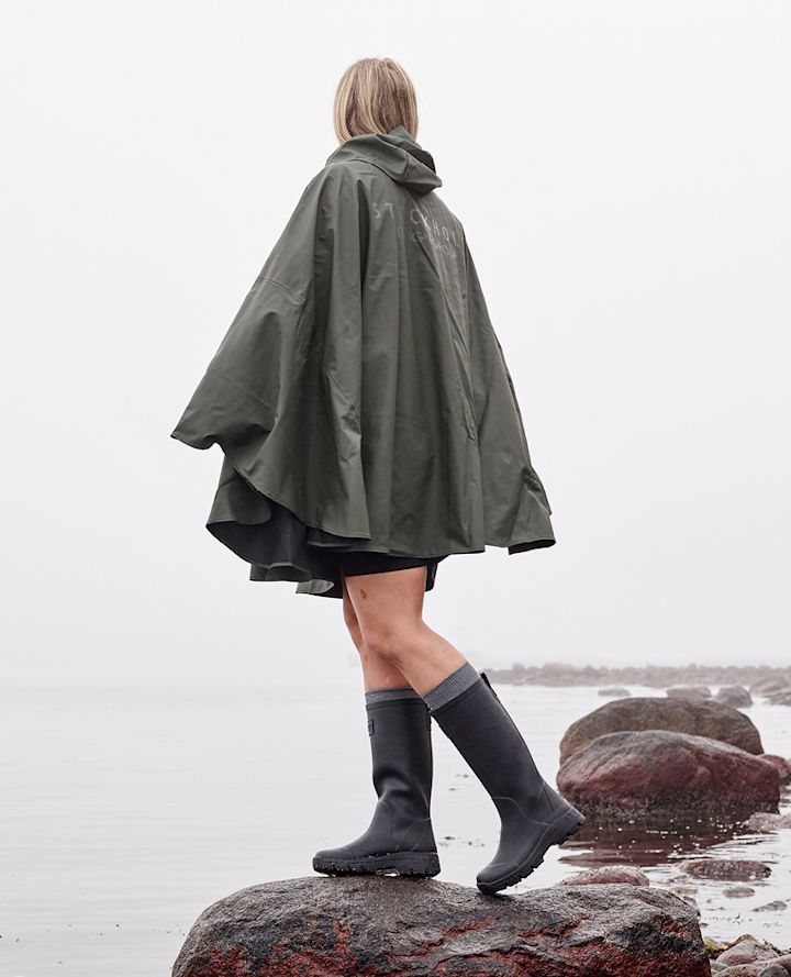 Person står på en stein ved vannet iført grønn regnponcho og svarte støvler på en tåkete dag.